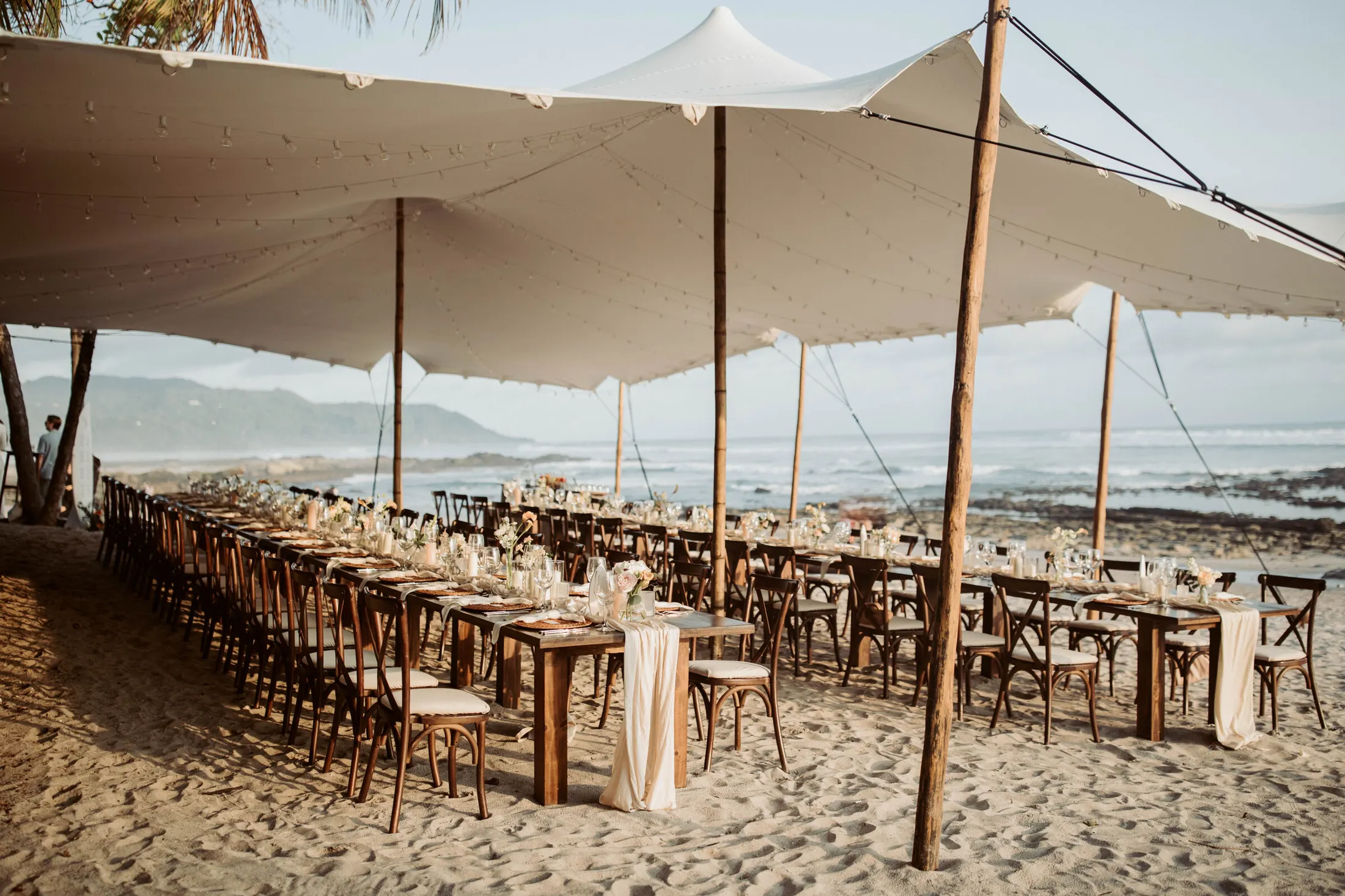 Long banquet tables set on the sand beneath a stretch tent hung with string lights, the surf breaking behind