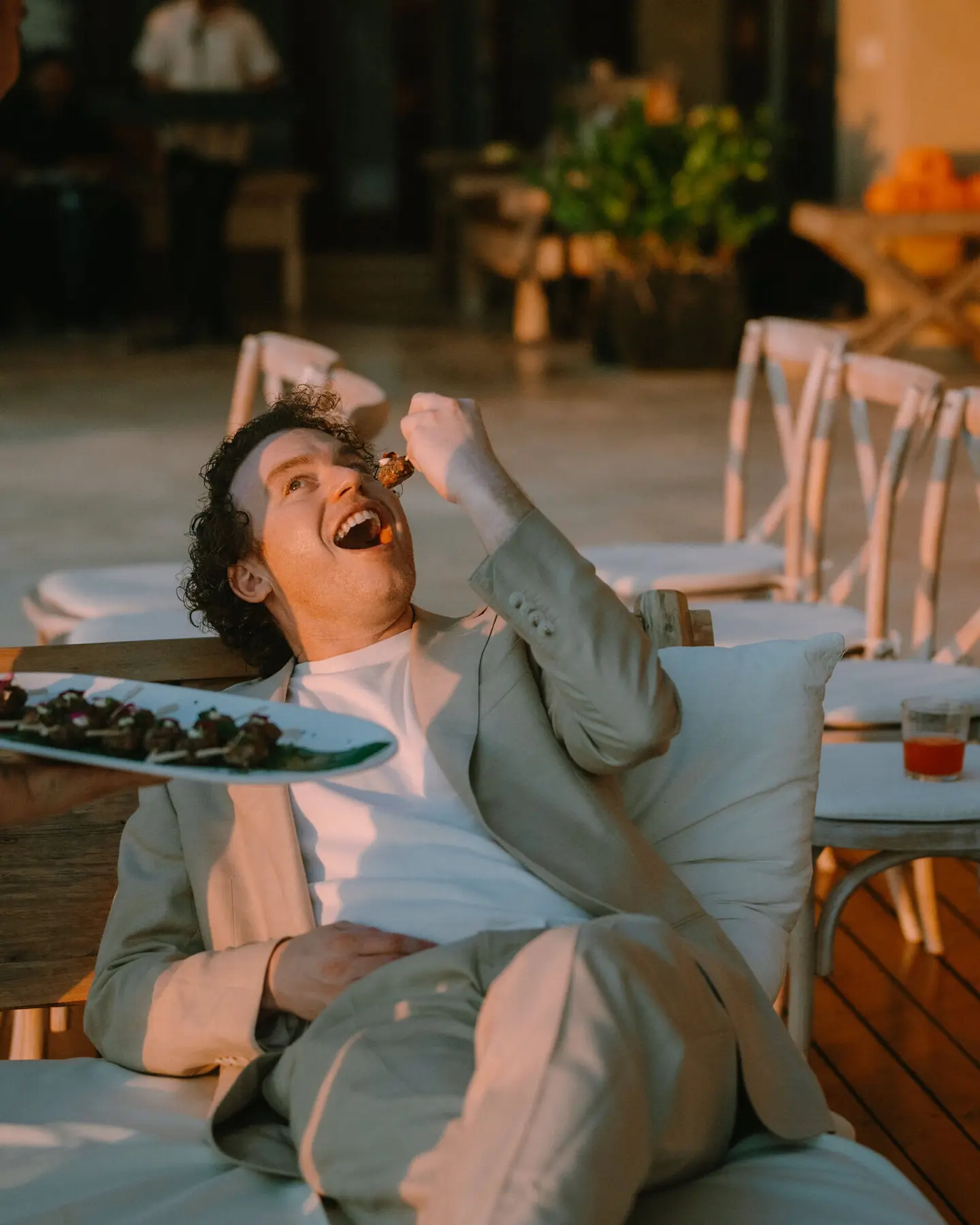 A guest stretched out on a daybed at golden hour, laughing as he takes a canapé from a platter held out beside him