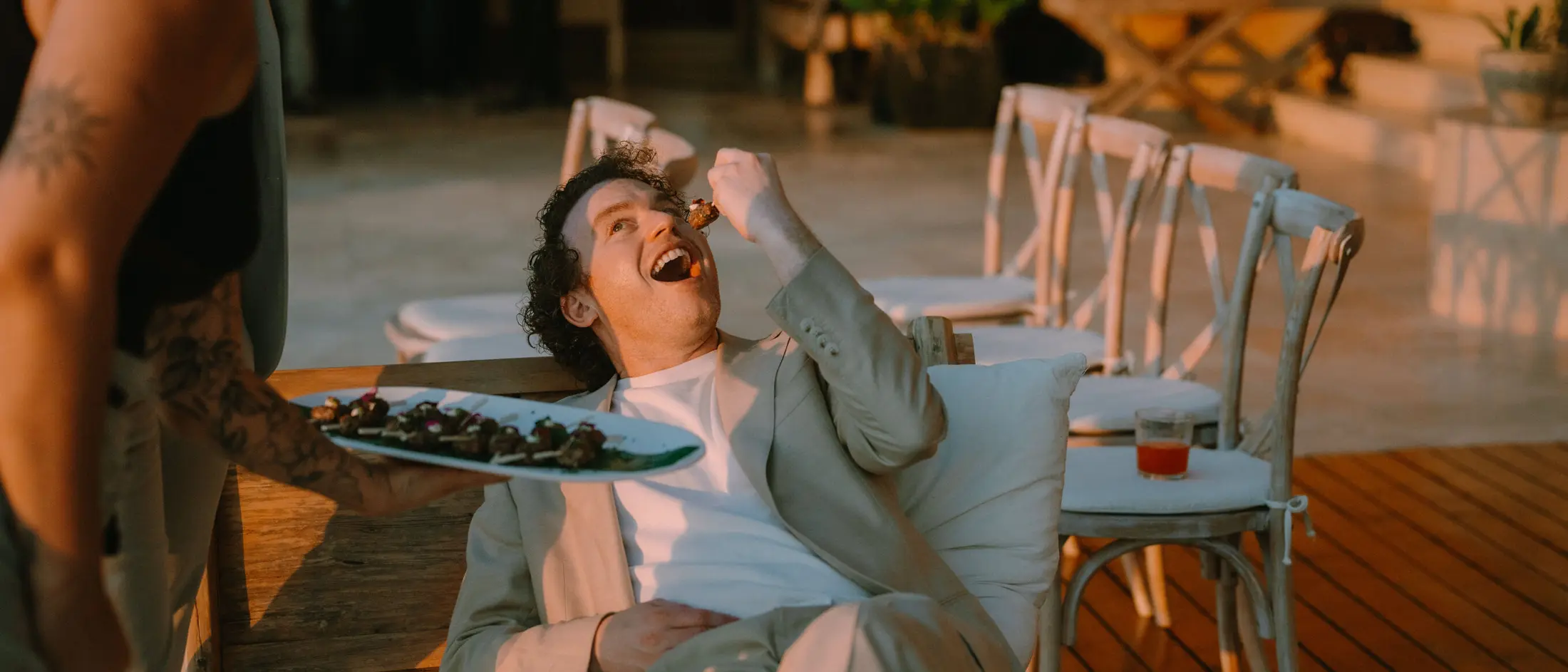 A guest stretched out on a daybed at golden hour, laughing as he takes a canapé from a platter held out beside him