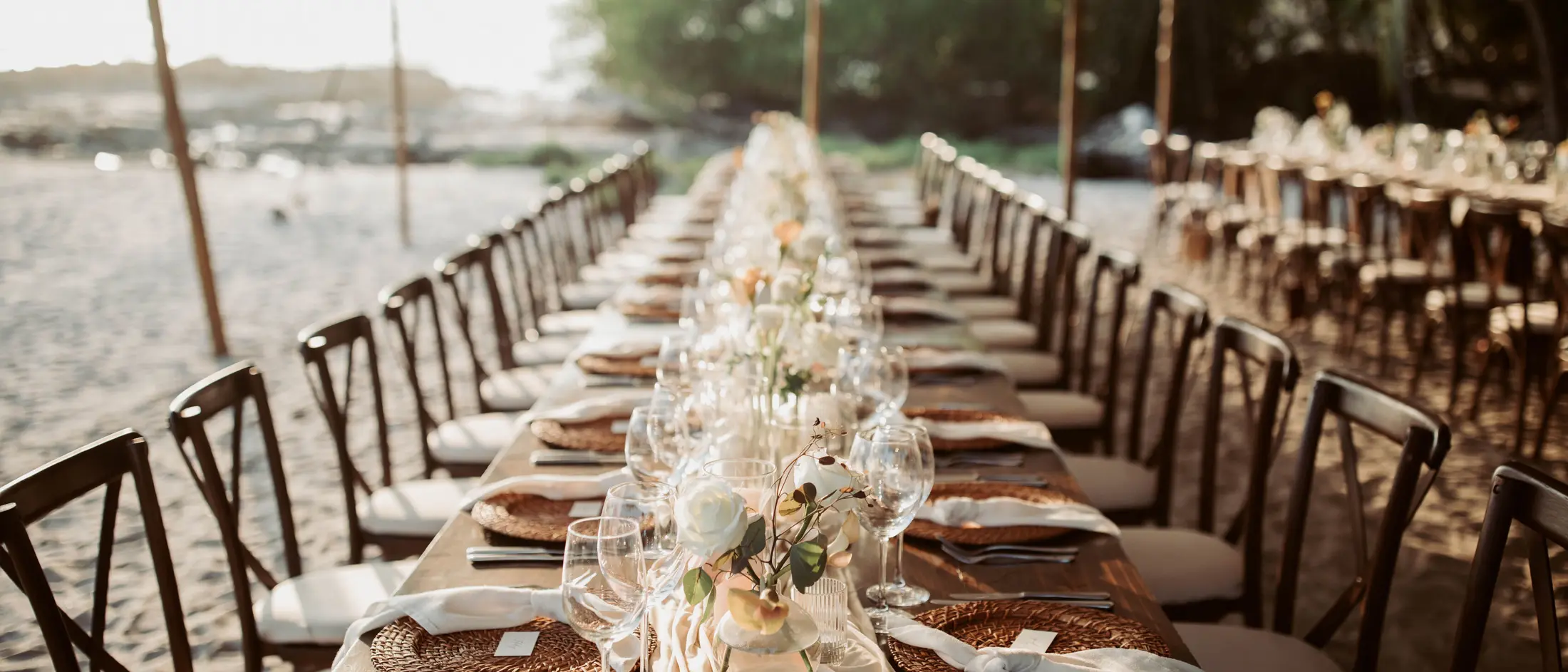 A long table laid on the sand under a stretch tent — woven chargers, linen runners and low garden roses receding down the table