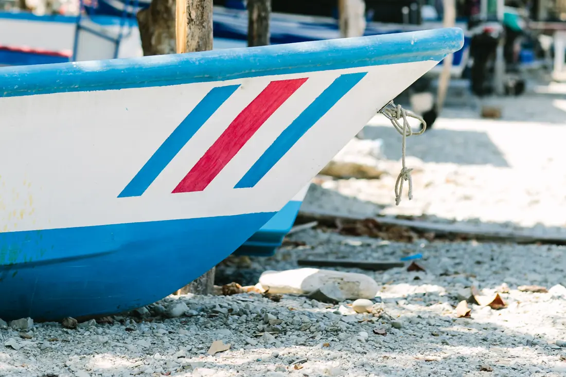 The painted bow of a fishing panga drawn up on a pebble beach beneath palapa shade