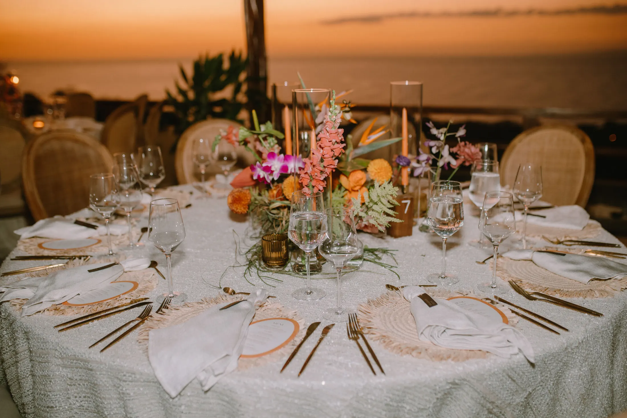 Table laid for dinner above the ocean at sunset — tapered candles, tropical florals and cane-backed chairs