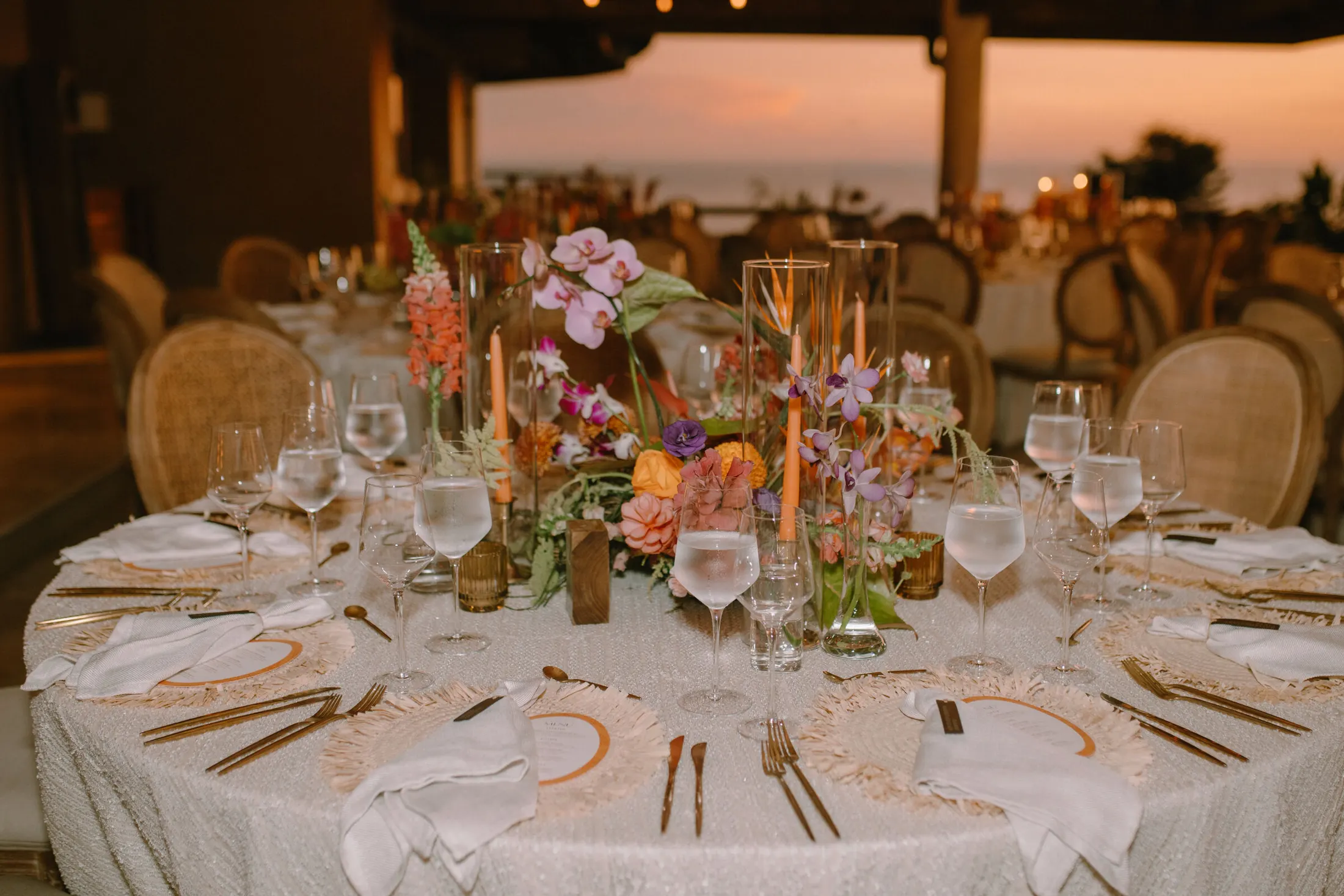 Round table laid for dinner at dusk, orchids and tapered candles at the centre, further tables and the ocean beyond