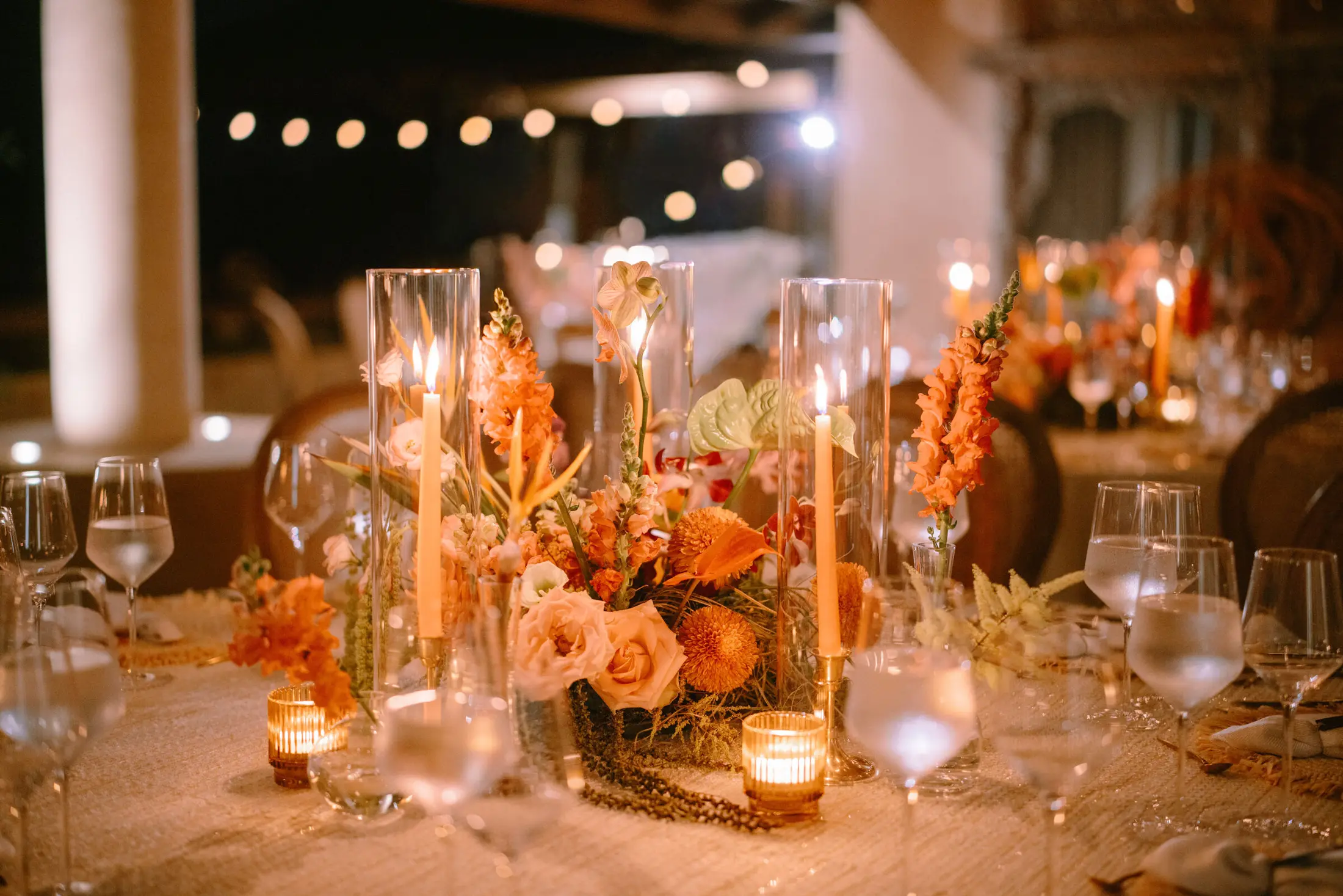 Reception table after dark — tapered candles in tall glass hurricanes, coral and orange florals, stemware and fringed placemats
